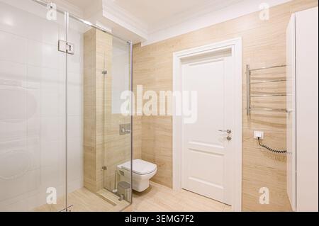 salle de bains bien éclairée avec cabine de douche en verre, toilettes blanches, carrelage à motifs en bois et porte blanche. La chambre a un esprit esthétique propre et moderne Banque D'Images