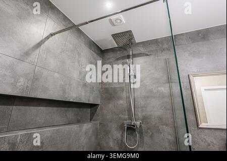 Douche moderne avec murs carrelés gris, pomme de douche carrée, panneau en verre et étagère intégrée. Banque D'Images
