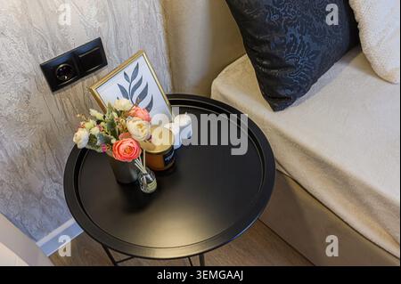 la table d'appoint noire contient des fleurs, une bougie et un cadre photo à côté d'un lit avec des oreillers. Banque D'Images