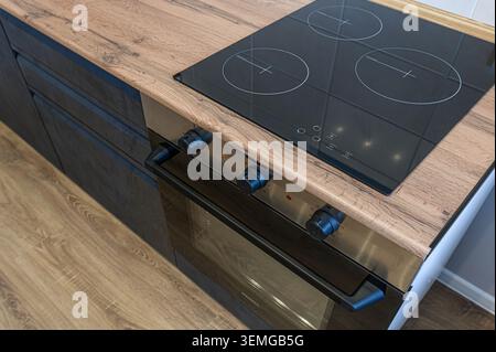 gros plan d'une cuisine moderne avec une table de cuisson noire élégante, un four avec boutons noirs et un comptoir en bois. La cuisine a des armoires sombres et Li Banque D'Images