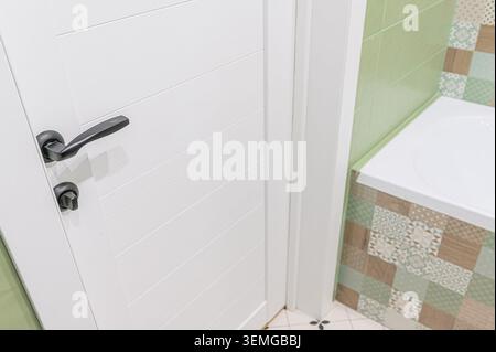 vue partielle sur une porte blanche avec une poignée noire, à côté d'une salle de bains avec carrelage vert et à motifs et une baignoire blanche. Banque D'Images