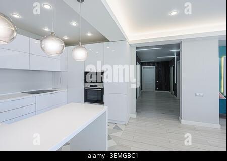 L'intérieur moderne dispose d'une cuisine blanche élégante avec des suspensions, des armoires brillantes et une porte ouverte menant à un couloir avec des murs gris et mult Banque D'Images