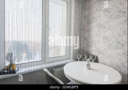chambre lumineuse et confortable avec une grande fenêtre, de délicats rideaux, une table blanche et d'élégantes chaises grises, le tout sur fond de papier peint à motifs. Banque D'Images