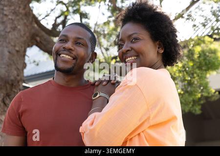 Couple afro-américain souriant, debout près de la maison sous l'arbre, montrant montre et bracelet Banque D'Images