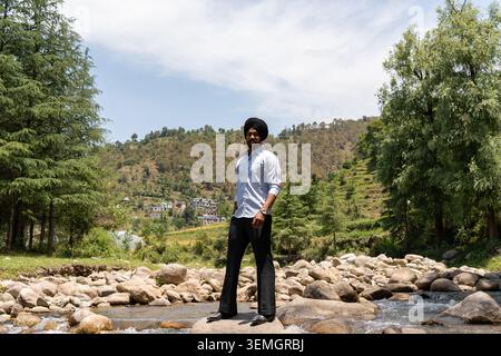 Un large portrait horizontal d'un jeune homme sikh en vêtements formels regardant la caméra debout sur des rochers dans une rivière de montagne. Banque D'Images