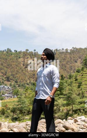 Un cliché vertical de trois quarts d'un jeune homme sikh vêtu de vêtements formels regardant loin avec confiance sur un magnifique fond montagnard. Banque D'Images