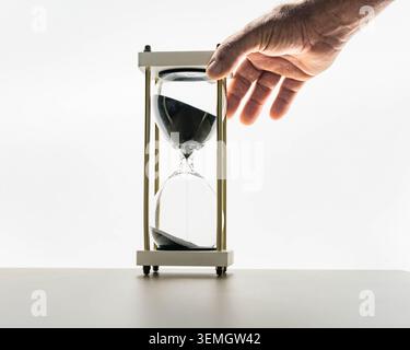 Main atteignant vers sablier de sable noir sur fond blanc, le temps qui passe et le concept de compte à rebours pour la date limite, la patience, la concentration et la productivité aujourd'hui Banque D'Images