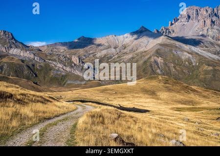 Un sentier de terre sinueux serpente à travers de vastes prairies de haute altitude aux tons dorés chauds, entourés d'imposantes chaînes de montagnes et de pics rocheux. La terre Banque D'Images