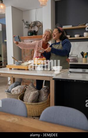 Diverses amies féminines portant tablier prenant selfie avec smartphone derrière l'îlot de cuisine avec gâteau Banque D'Images