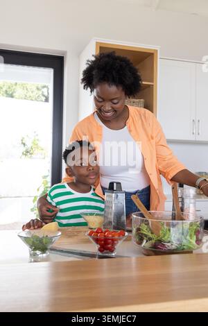 Mère afro-américaine et fils d'âge scolaire préparant la salade dans la cuisine à l'île en bois avec râpe Banque D'Images