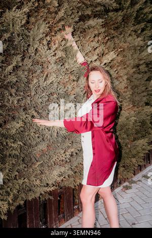 Femme haie mode femme élégante portant une robe blanche et chemise rouge posant contre un grand mur à feuilles persistantes dans un jardin Banque D'Images