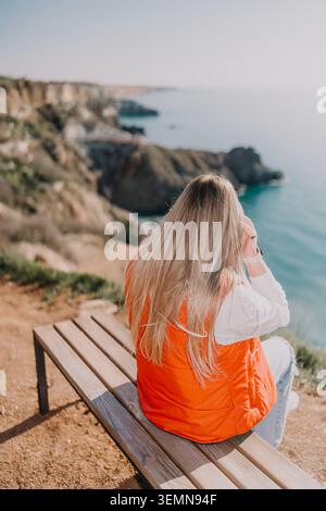 Femme falaise mer assis sur un banc en bois surplombant une côte pittoresque et des formations rocheuses accidentées au cours d'une aventure de voyage paisible Banque D'Images