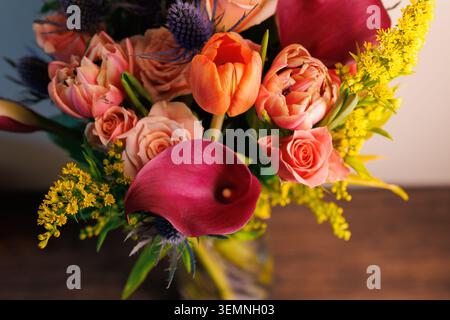 Un bouquet vibrant de diverses fleurs est disposé dans un vase clair sur une table en bois. Les fleurs montrent différentes formes, couleurs et tailles sous lumineux Banque D'Images