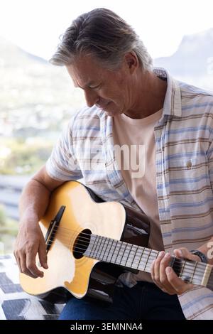 Homme mûr jouant de la guitare acoustique sur le balcon avec vue sur les collines de la ville, portant chemise rayée et montre Banque D'Images