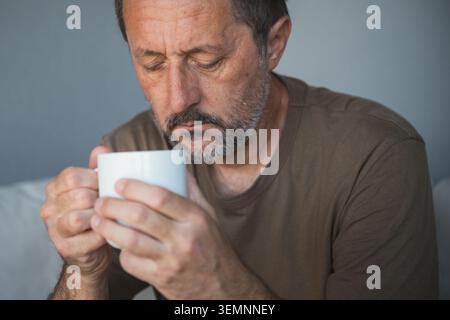 Homme d'âge mûr réfléchi avec la barbe tenant une tasse de café blanc regardant pensif à la maison, mise au point sélective Banque D'Images