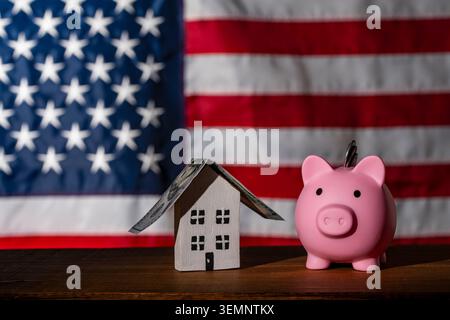 Tirelire et maison de jouet avec toit dollar sur fond de drapeau américain. Tirelire rose avec des pièces de monnaie à côté d'une petite maison en bois avec un toit de billet d'un dollar à nouveau Banque D'Images