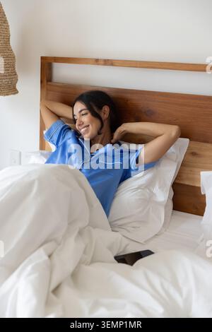 Femme en pyjama en satin haut étirant les bras derrière la tête dans le lit avec literie blanche et smartphone Banque D'Images