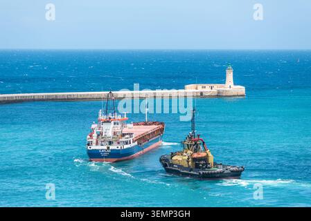Valletta, Malte – 16 mai 2025 : cargo et remorqueur naviguant près de l'entrée du port historique et du phare de la Valette, Malte, entouré de brick Banque D'Images