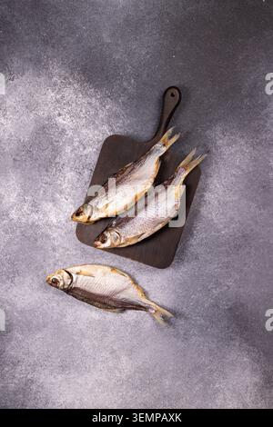 Poisson séché salé, une collation traditionnelle, reposant sur une planche de bois sombre et directement sur un fond gris texturé, symbolisant le prépar alimentaire traditionnel Banque D'Images