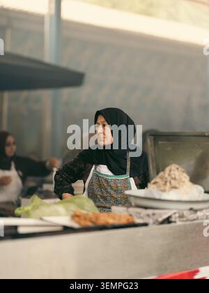 TACHKENT, OUZBÉKISTAN – JUIN 2025 : une vendeuse ouzbèke âgée dans un hijab noir traditionnel debout à son stand de nourriture de rue au bazar de Chorsu. Banque D'Images