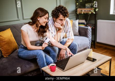 Un jeune couple est assis ensemble sur un canapé, comparant les options sur un ordinateur portable tout en faisant un achat en ligne. La femme détient une carte de crédit et pointe à la Banque D'Images