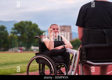 Athlète féminine en fauteuil roulant souriante se relaxant sur la piste après l'entraînement avec son coéquipière au coucher du soleil Banque D'Images