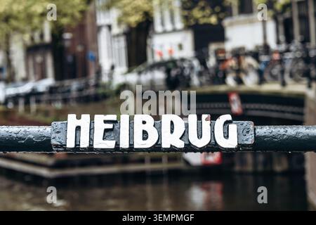 Vue rapprochée du panneau de nom du pont Heibrug au-dessus d'un canal d'Amsterdam, avec l'eau, les vélos et les environs urbains historiques légèrement flous à l'arrière Banque D'Images