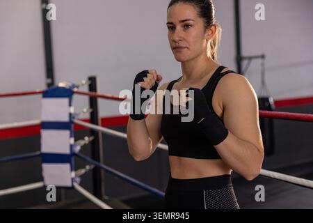 Boxeur féminin debout dans l'anneau de boxe, soulevant les poings enveloppés dans les vêtements de sport, formation avec sac lourd Banque D'Images