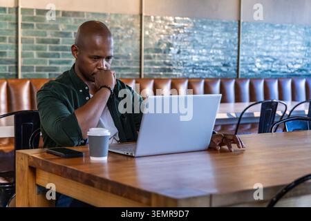 Homme afro-américain assis au café dans la chemise vert foncé tapant sur ordinateur portable argenté avec du café Banque D'Images