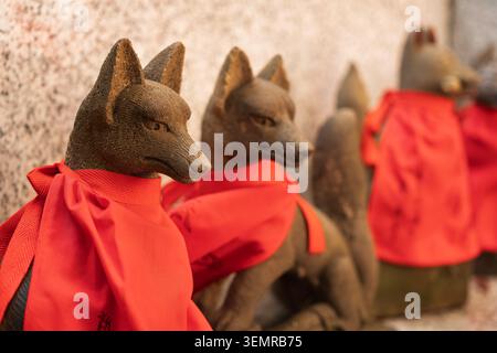 Rangée de plusieurs petites statues de renard en pierre portant des bavoirs en tissu rouge alignés dans un sanctuaire Inari, Tokyo, Japon Banque D'Images