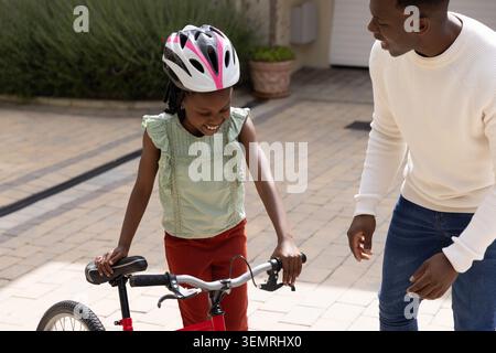 Père afro-américain et fille pratiquant le vélo rouge sur l'allée pavée avec casque Banque D'Images