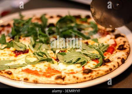 Pizza artisanale fraîchement cuite garnie de roquette et de mozzarella fondue tandis qu'un coupe-pizza se prépare à servir une tranche. Banque D'Images