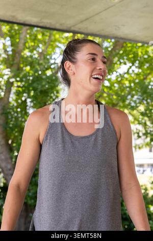 Femme debout sous le surplomb de béton portant un débardeur gris, cheveux dans le chignon, rire au milieu des arbres Banque D'Images
