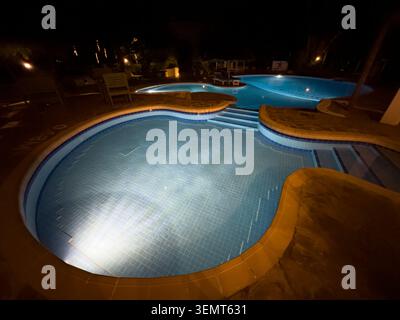 Une piscine de luxe à la forme libre brille avec un éclairage sous-marin la nuit dans un hôtel de villégiature tropical haut de gamme, créant une ambiance étonnante. Banque D'Images