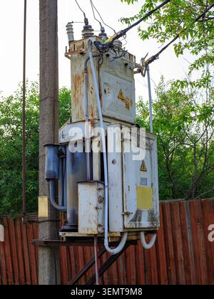 Vieux transformateur de puissance rouillé est monté sur un poteau en béton près d'une clôture en bois brun. Infrastructure rurale de distribution d'électricité à haute tension c Banque D'Images