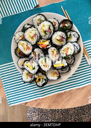 Les rouleaux de sushi sont disposés soigneusement sur une assiette blanche. Les rouleaux contiennent du concombre, du poisson rouge et des œufs. Ils sont servis avec des baguettes sur un tapis bleu et vert Banque D'Images