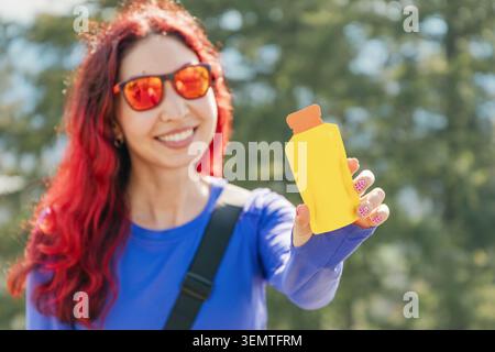 Femme souriante, tenant une boisson gel snack jaune, favorisant un mode de vie sain et une nutrition rapide en plein air Banque D'Images