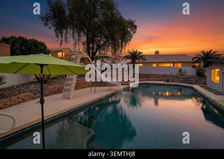 Une scène tranquille dans la cour arrière au coucher du soleil avec une piscine rectangulaire, un toboggan incurvé coulissant et un parasol vert vif. Lumière chaude du soir Banque D'Images