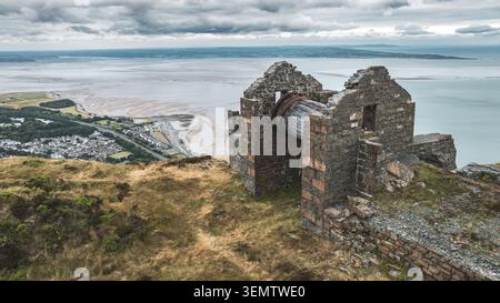 Un bâtiment en pierre abandonné avec une grande roue en bois se dresse sur une colline herbeuse au-dessus d'une ville balnéaire et un large estuaire sablonneux sous un ciel nuageux Banque D'Images