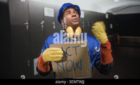 Homme constructeur tenant un panneau en carton lisant agir maintenant et faisant des gestes vers le haut dans un vestiaire d'un bâtiment tout en portant un casque et des gants ; plaidoyer urgent. Banque D'Images