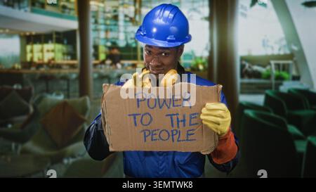 Homme ouvrier de la construction tenant le panneau en carton lisant le pouvoir aux gens, portant un casque, des protections auditives et des gants tout en souriant légèrement dans un bâtiment Banque D'Images