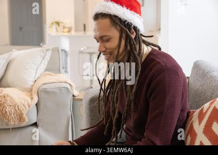 Homme afro-américain assis sur le canapé gris dans le salon portant chapeau de père Noël pull marron Banque D'Images