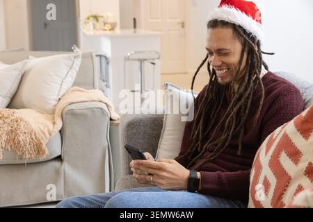 Homme assis sur le canapé gris tenant smartphone, portant chapeau de Père Noël, pull Bordeaux, espace de copie Banque D'Images
