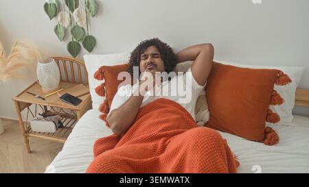 Homme allongé sur le lit avec une couverture orange serrant la poitrine et la main sur la gorge à côté de la table de chevet et smartphone dans le studio ; repos inconfortable. Banque D'Images