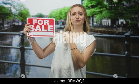 La femme pointe le doigt à pour le signe de vente sur la rampe de canal de rue tout en tenant l'écriteau blanc et souriant ; promotion de la confiance des ventes. Banque D'Images