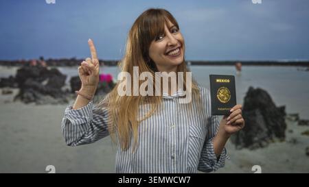 Femme souriante tient le passeport mexicain dans la main droite et pointe l'index vers le haut tout en se tenant debout sur une plage de sable avec des roches volcaniques et l'horizon de la mer ; trav Banque D'Images