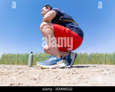 Profil à faible angle d'un homme caucasien d'âge moyen squattant pour se reposer sur le sentier de terre à côté du champ de culture verte pendant la canicule estivale Banque D'Images
