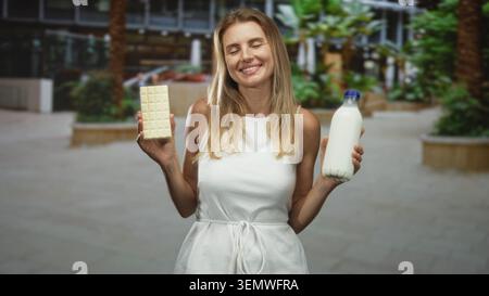 Femme tient une barre de chocolat blanc et une bouteille de lait avec les yeux fermés et souriant dans l'atrium du bâtiment ; plaisir de contentement. Banque D'Images