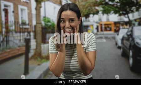 Femme touchant les joues avec les mains, souriant et regardant agréablement surpris dans la rue avec des voitures garées et des devantures ; célébration de joie. Banque D'Images