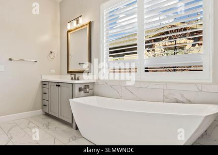 La salle de bains blanche et grise aux couleurs vives est dotée d'une baignoire ovale autoportante, d'un sol en marbre et d'un meuble-lavabo gris élégant avec un meuble à miroirs. Une grande fenêtre avec whi Banque D'Images